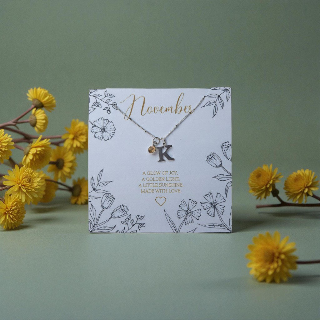 Personalised November citrine birthstone and initial necklace displayed on a floral illustrated card reading ‘A glow of joy, a golden light, a little sunshine. Made with love.’ Surrounded by yellow flowers on a soft green background.