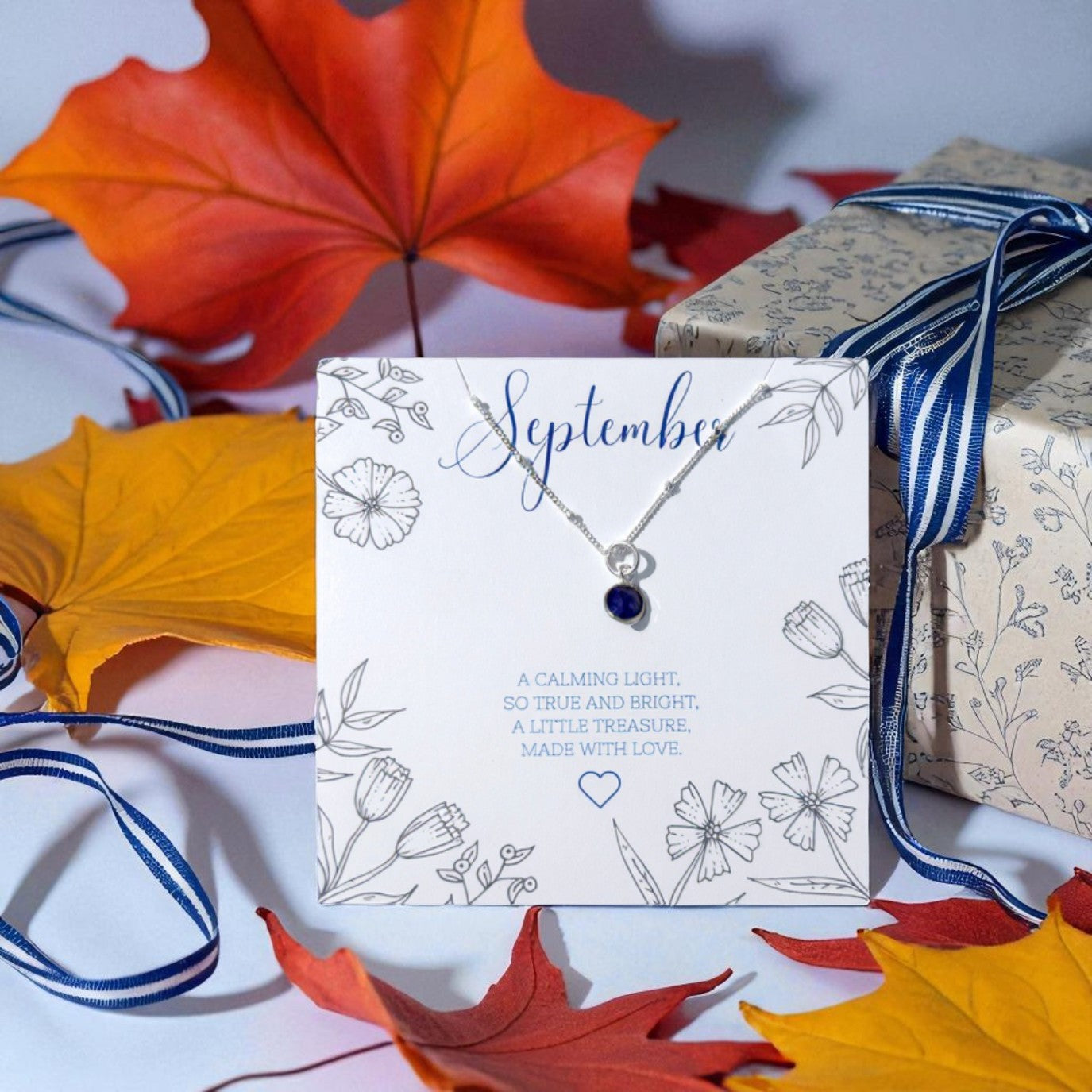 Made Here With Love September birthstone necklace featuring sapphire pendant on sterling silver chain, displayed on illustrated message card with autumn leaves and gift box, meaningful handmade birthday jewellery gift UK