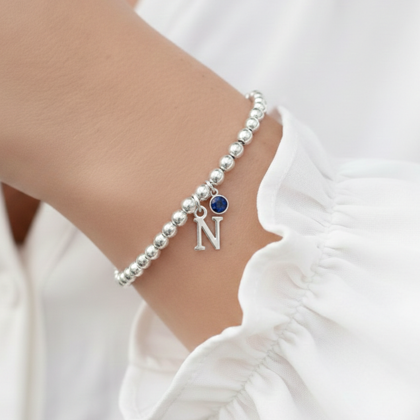 Silver beaded bracelet with a blue gemstone and letter 'N' charm on a wrist against a white background.