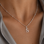 A close-up of a delicate silver chain necklace worn on a model's neck, featuring a small lowercase letter 's' pendant, styled with a satin gray shirt.
