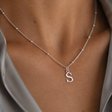A close-up of a delicate silver chain necklace worn on a model's neck, featuring a small lowercase letter 's' pendant, styled with a satin gray shirt.
