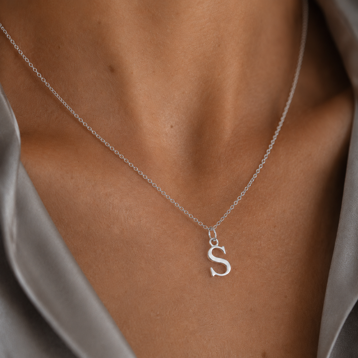 A close-up of a delicate silver chain necklace worn on a model's neck, featuring a small, polished silver initial 'S' pendant. The model is wearing a satin gray top.